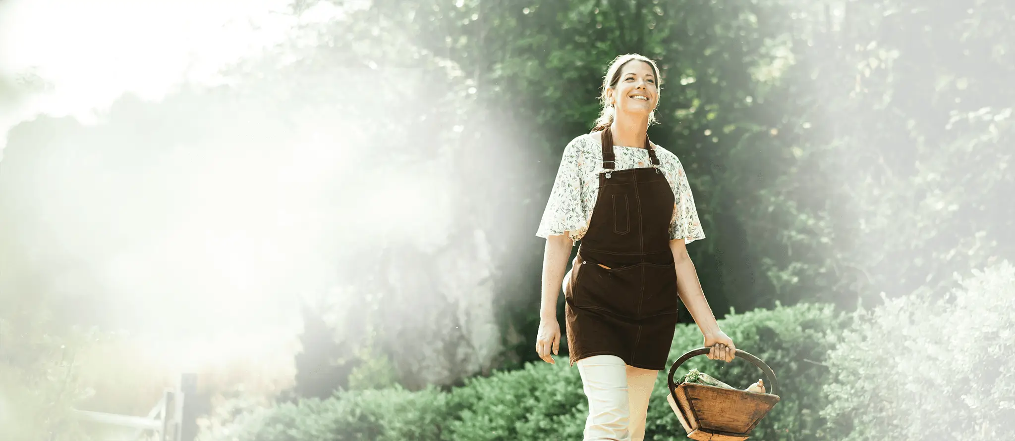 A smiling woman wearing gardening coveralls walks outside, holding a basket.